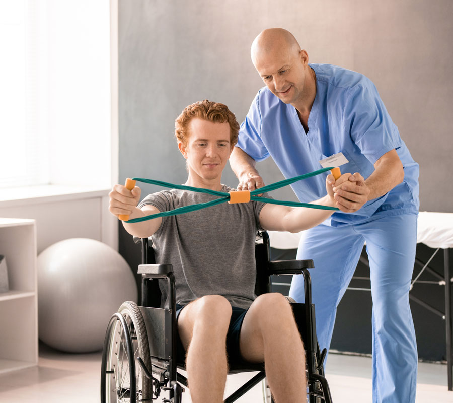Mature clinician in blue uniform supporting hand of young patient in wheelchair Mature clinician in blue uniform helping young male patient in wheelchair while supporting his left hand during physical exercise with resistance band